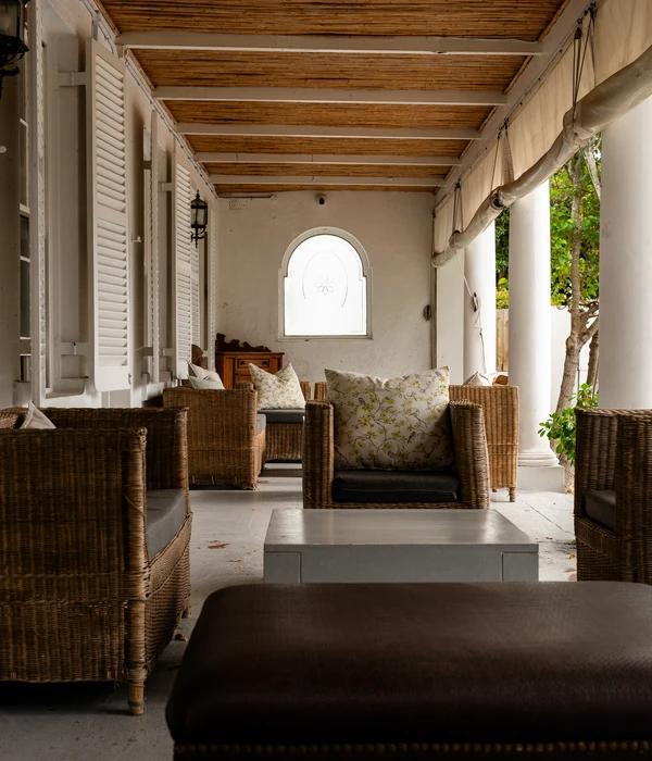 Een lang overdekt terras met rieten fauteuils, bloemenkussens, witte pilaren en een plafond in bamboestijl.