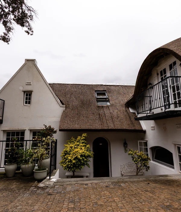 Binnenplaats van een wit landhuis met rieten daken, een geplaveide vloer, potplanten en een kleine boogvormige deuropening.