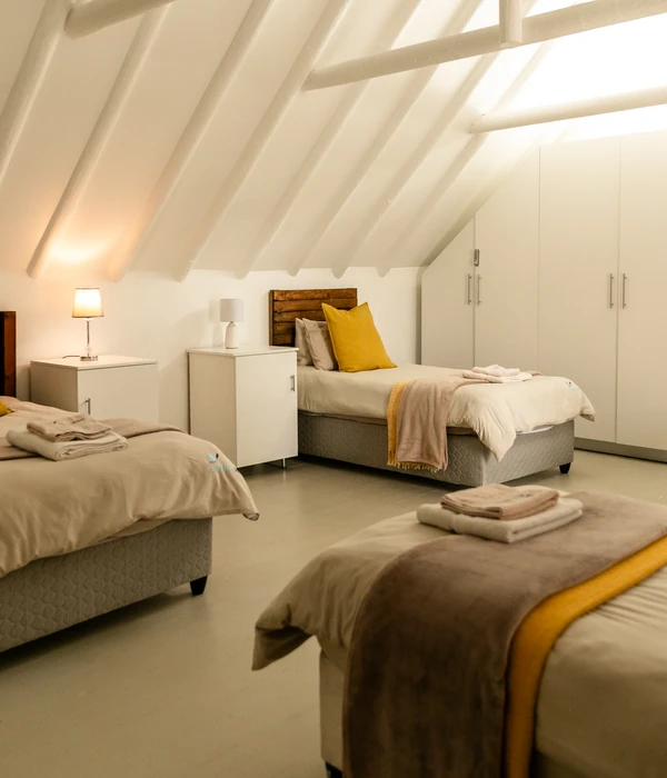 Een gedeelde zolderkamer met drie netjes opgemaakte bedden, gele accenten en witte kledingkasten onder een gewelfd plafond met zichtbare balken.