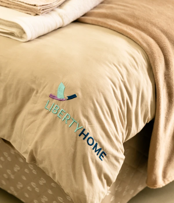 Close-up van een netjes opgemaakt bed met beige beddengoed, voorzien van een geborduurd "LIBERTY HOME"-logo, en opgevouwen handdoeken.