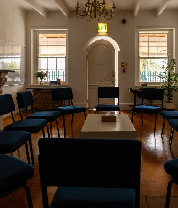 Een zonnige groepstherapieruimte met blauwe stoelen in een cirkel rond een lage tafel met tissues op een houten vloer.