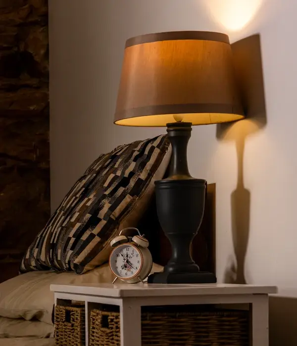Een warm nachtlampje gloeit op een wit nachtkastje naast een vintage wekker en een kussen met een patroon.