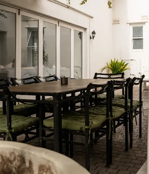Een houten eettafel voor buiten met zwarte metalen stoelen en groene kussens, geplaatst in een lichte, rustige binnenplaats.