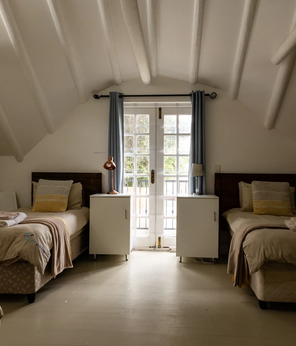 Lichte kamer met twee bedden, witte balken aan het plafond en openslaande deuren naar een balkon met blauwe gordijnen.